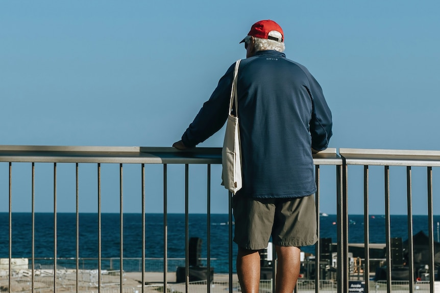 an Older man seen from the back stands at a railing and looks out to sea.