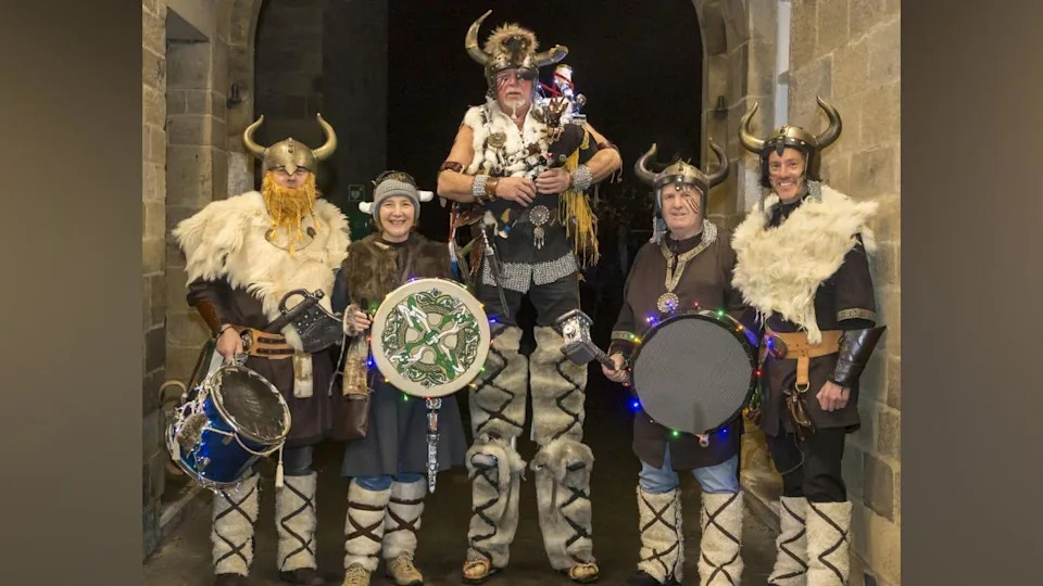Five of the Vikings stand in the hallways of an old stone building. They are wearing various Viking accessories including helmets with horns, fur leggings and leather arm braces. The man in middle is on stilts, but dressed in a way that makes him look like a very tall Viking. There are holding various instruments including horns, pipes and drums.