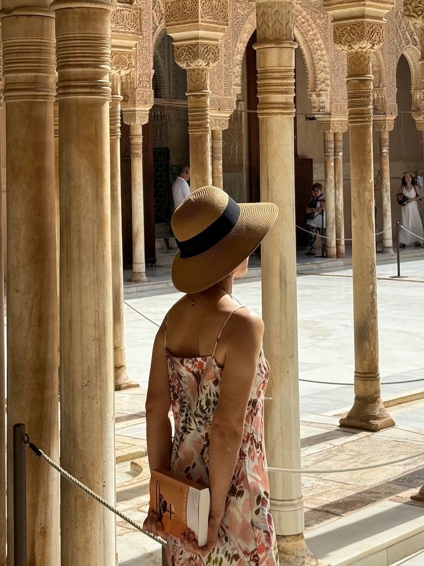A Woman in a sundress stands among stone pillars at Spanish tourism site. Her faces is hidden by a straw hat.