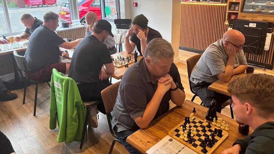 A group of men play chess on small square tables in a cafe.