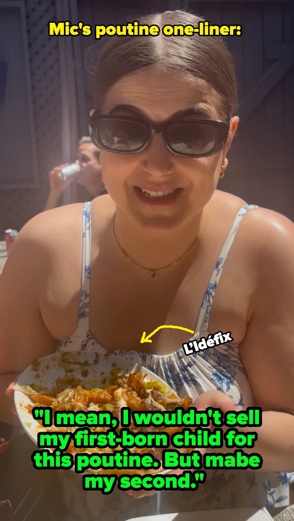 Person with short hair sitting outdoors, holding a plate of food, smiling gladly at it. People are seated at tables in the background
