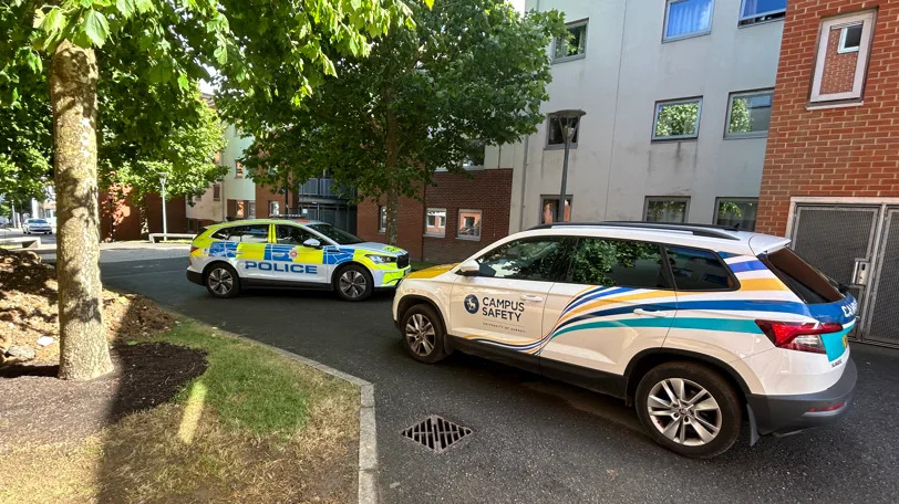 Two cars are parked up outside a building. It is sunny and there is a big tree. 