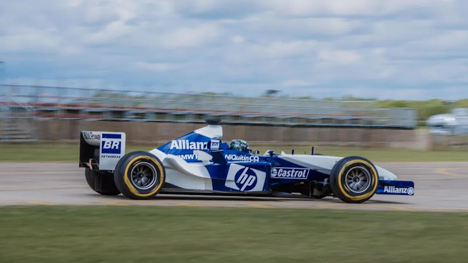 A restored FW25 F1 car. It is white and blue and is travelling around a corner at a track.