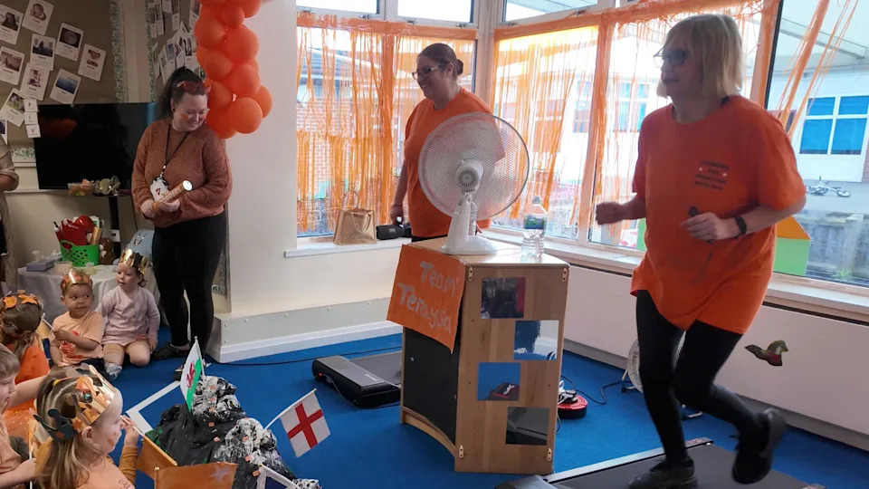 Two women in orange t-shirts running on black treadmills as children look on.