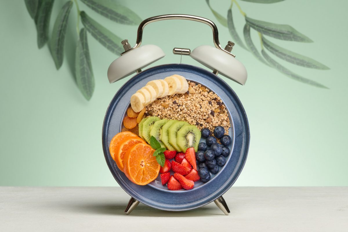 Alarm clock with a dial replaced by a plate full of a healthy breakfast. The plate contains granola, banana slices, kiwi, tangerine, strawberries, and blueberries. Green background