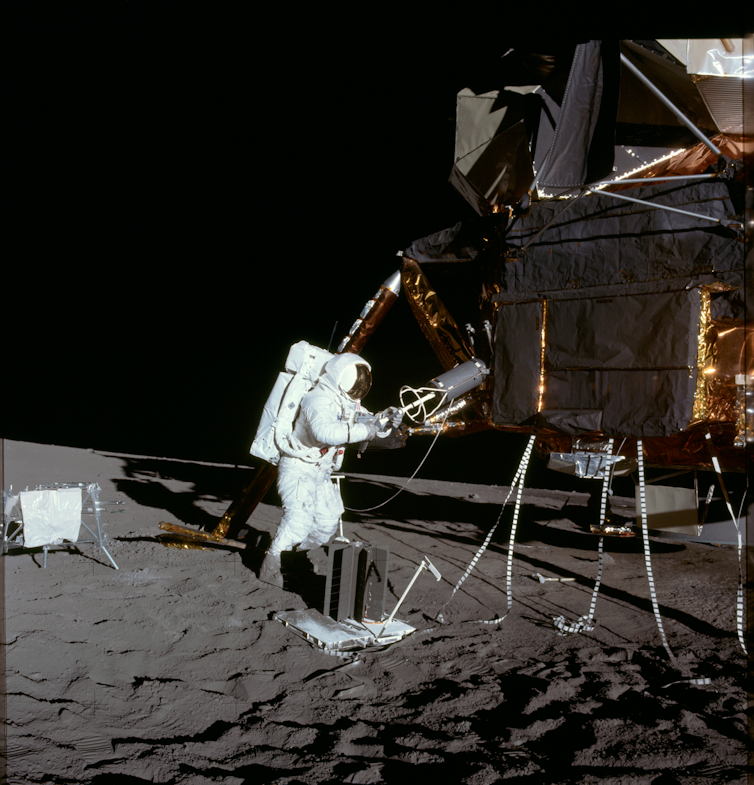 An astronaut in a white spacesuit tending to machines on a grey, dusty moon.