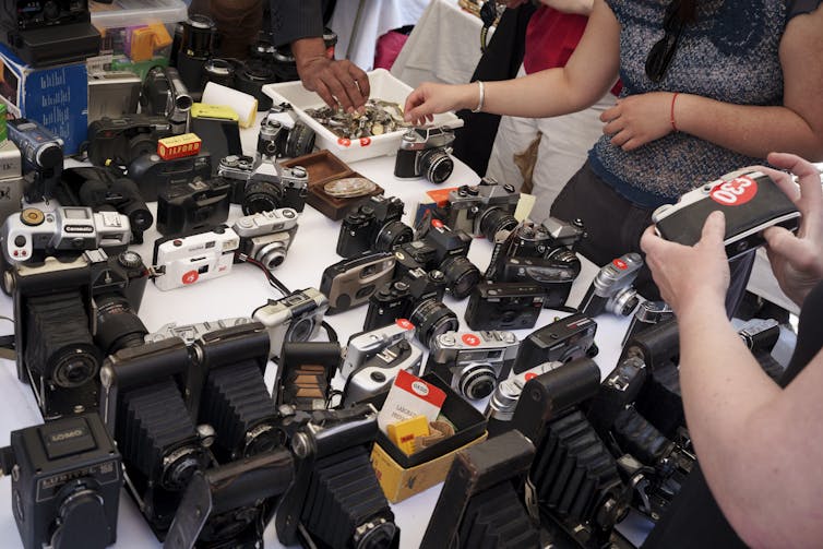 A table featuring an array of camera equipment spanning different eras, with hands holding some of the objects.