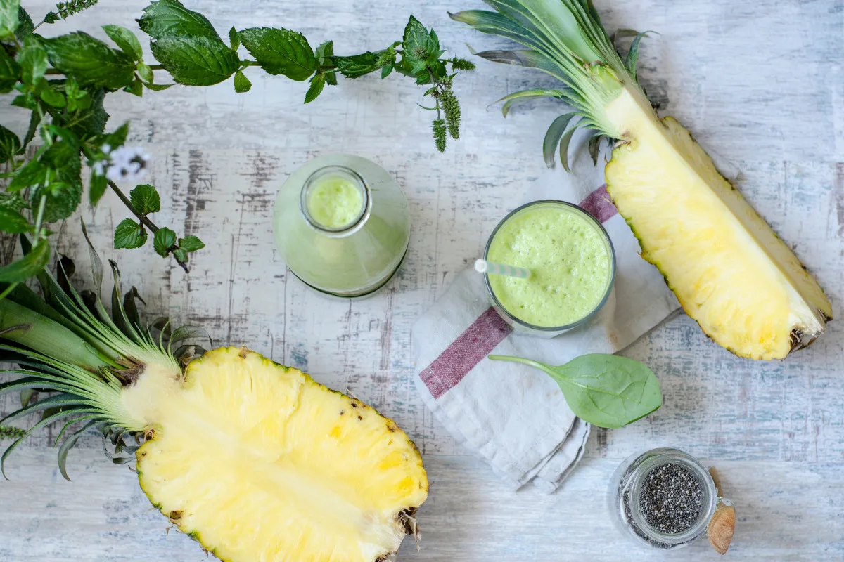 Top down image of a pineapple cut in two and a a glass of green healthy drink, spinach and mint can also be seen