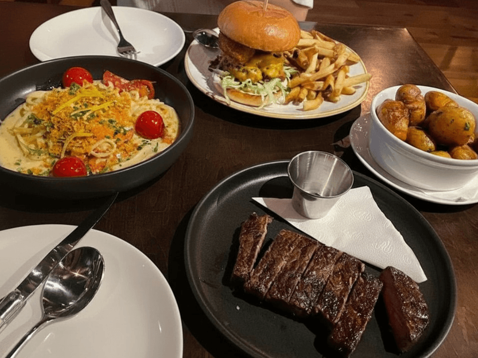 Food spread at The Whalesbone with sliced steak, a burger with fries, a bowl of pasta, and a side of potatoes on a restaurant table.