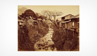 Sepia-toned photo of a narrow rocky stream flowing between traditional wooden houses on stilts, with bare trees and mountains visible in the background.