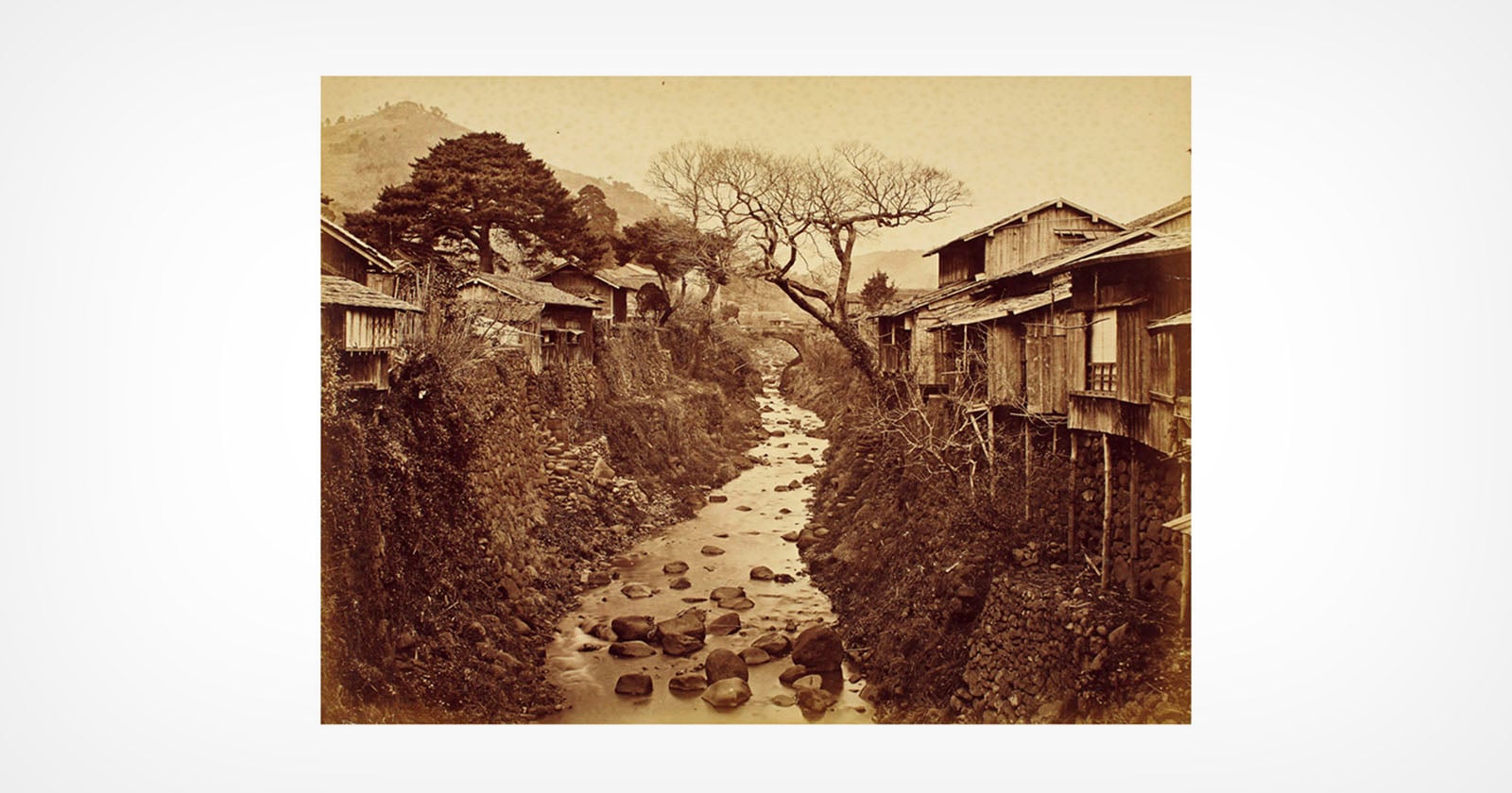 Sepia-toned photo of a narrow rocky stream flowing between traditional wooden houses on stilts, with bare trees and mountains visible in the background.