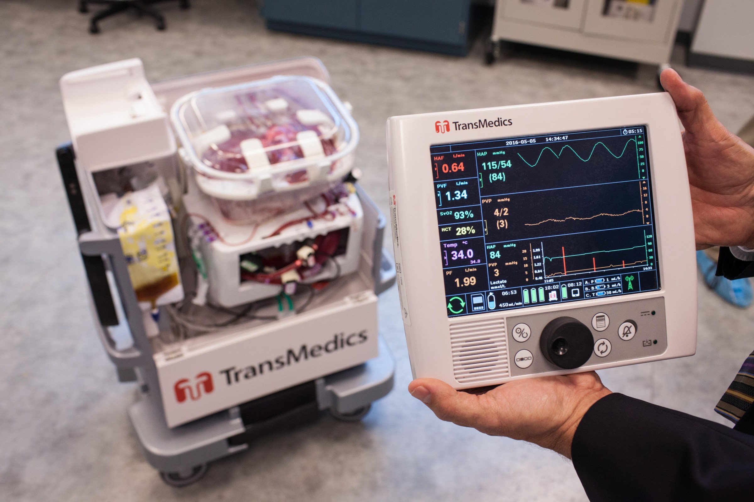 A man’s hands hold an electronic medical device in the foreground while a robotic cart sits in the background
