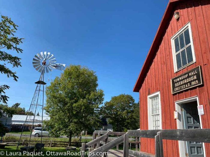 two-storey building with reddish siding, with a split-rail fence in foreground and windmill in background
