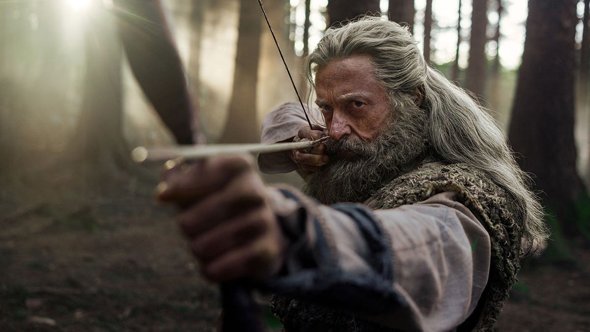 Hugh Jackman as a long-haired man drawing a bow and arrow in a forest.