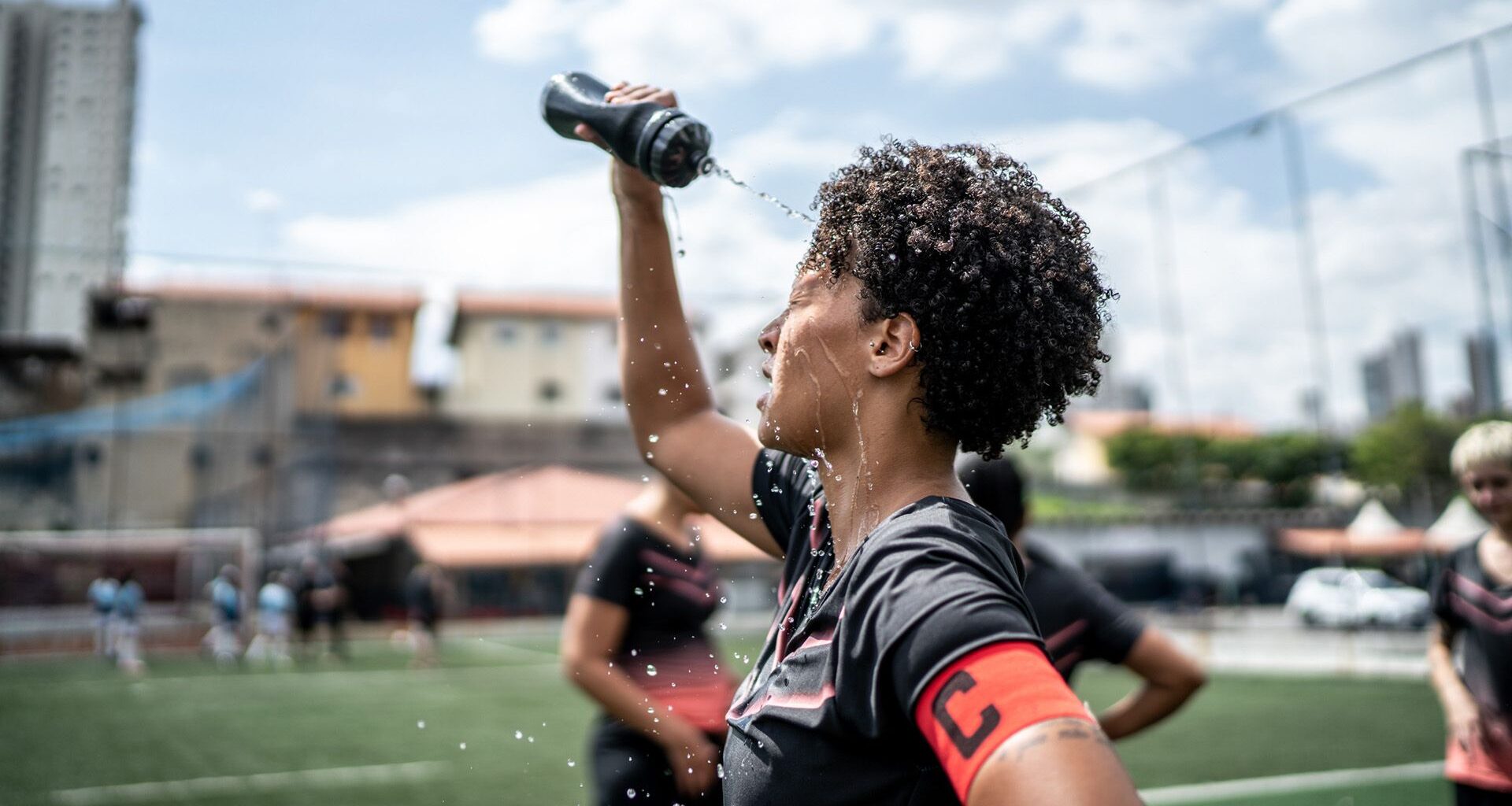 A woman with short, dark curly hair sprays herself with a water bottle standing on a grass turf.
