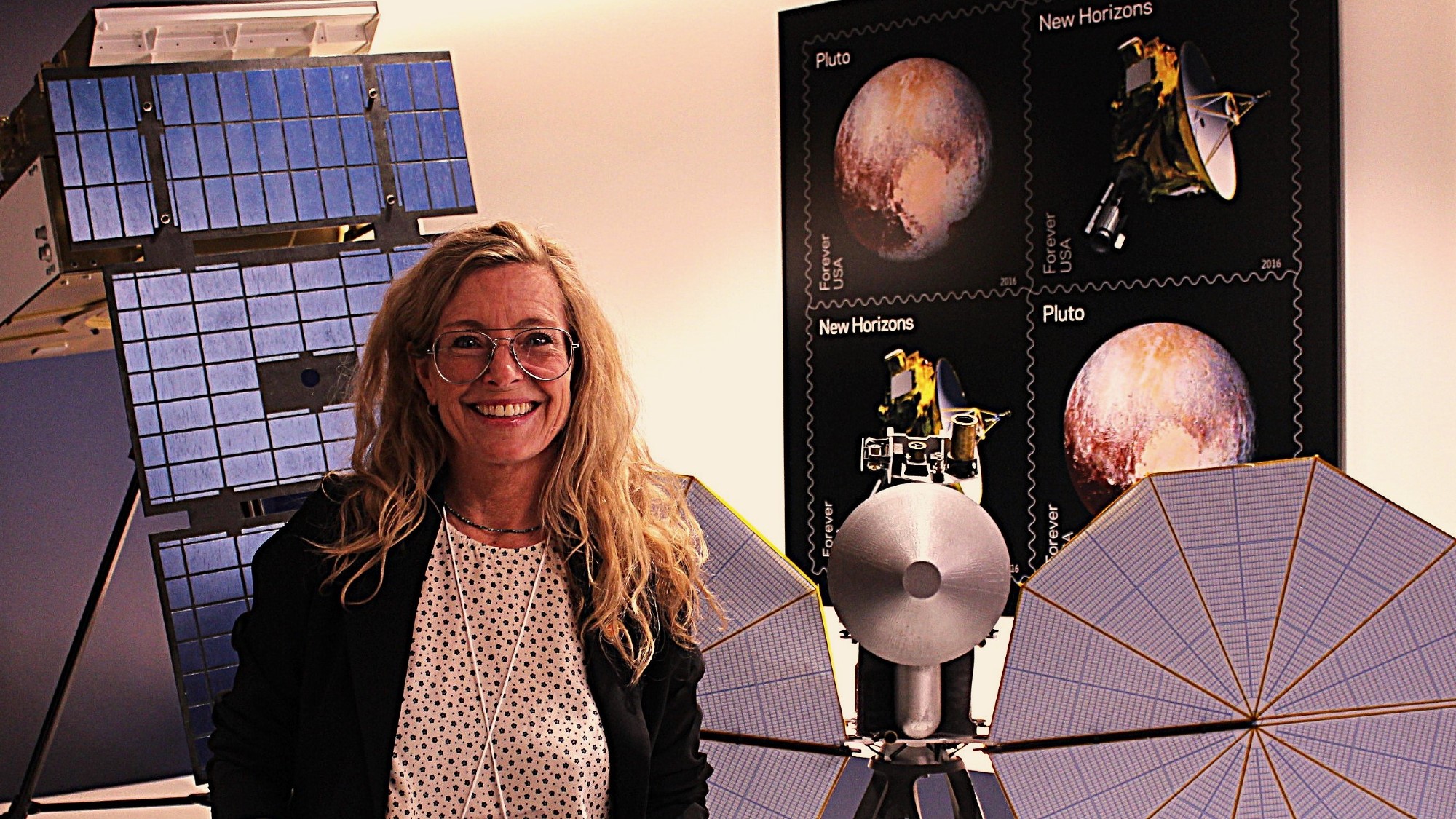 a woman wearing glasses smiles in front of a model of a spacecraft with two large octagonal solar arrays