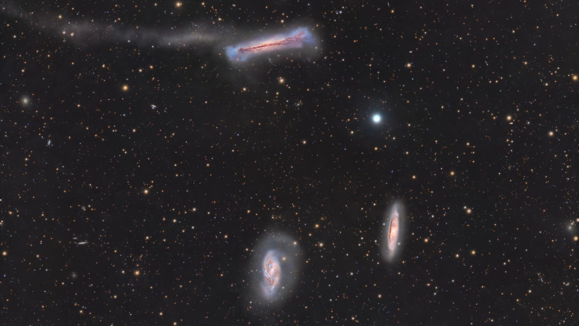 A trio of galaxies are pictured shining in deep space against a starfield dotted with more distant galaxies.