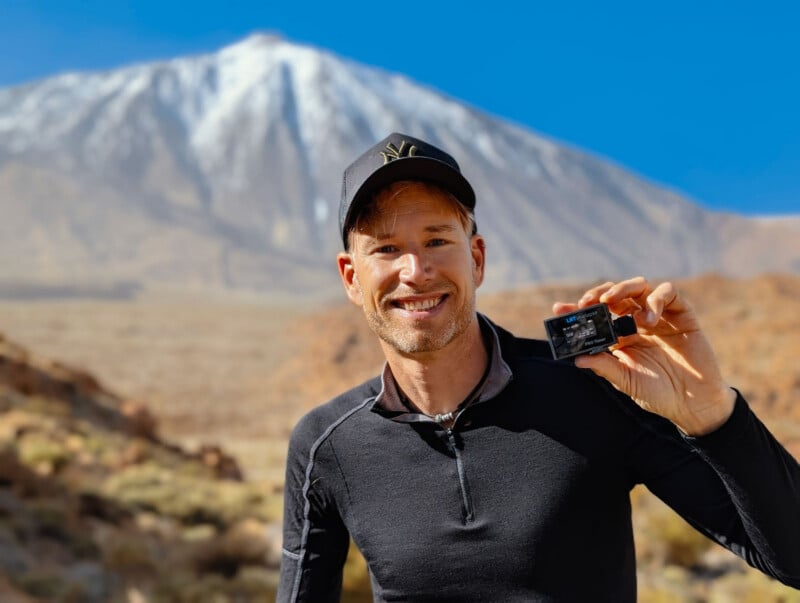 A smiling man in a black shirt and cap stands outdoors, holding a small black device. Behind him is a landscape with dry terrain and a tall, snow-capped mountain under a clear blue sky.