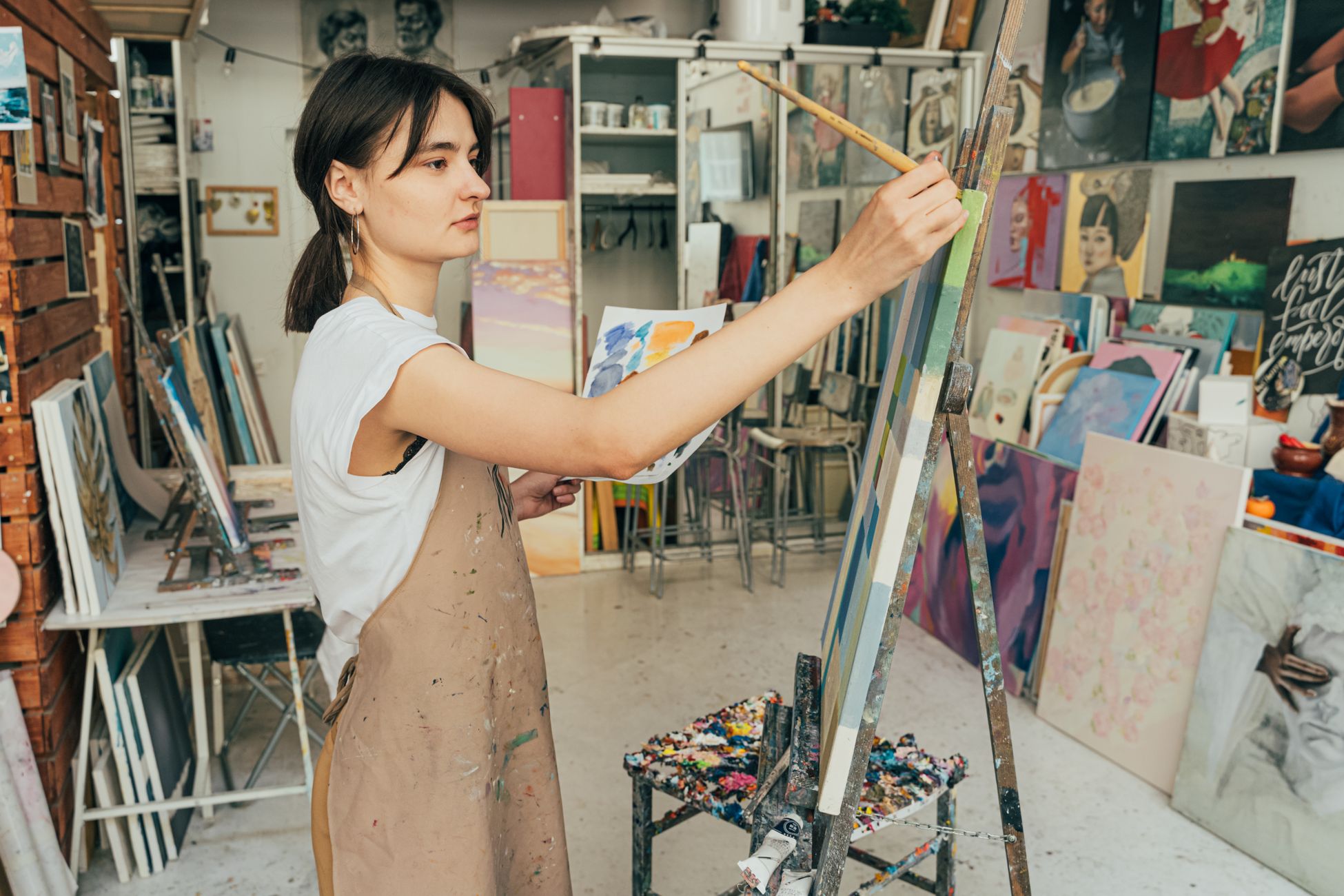 A young female artist painting on a canvas in an art studio filled with creative works.