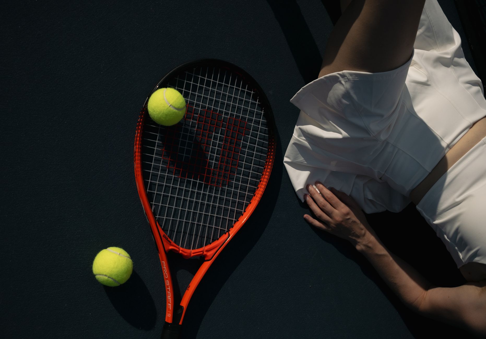 Tennis player in white sportswear next to racket and tennis balls on court.
