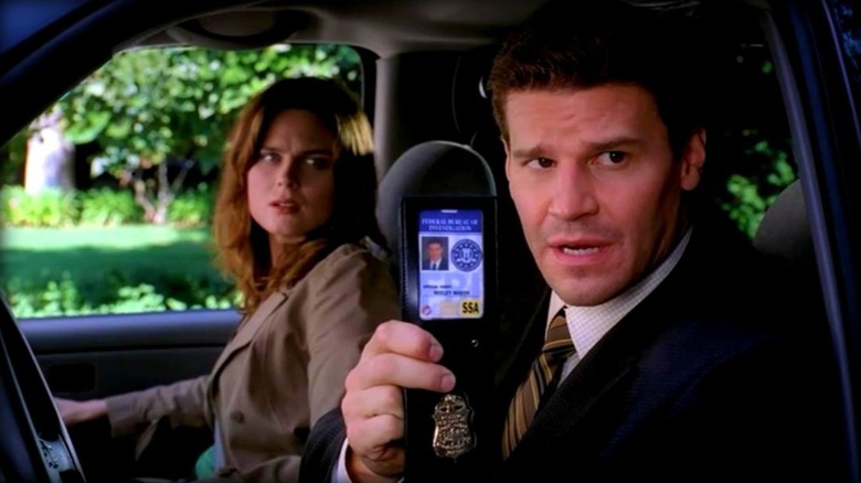 Booth holding up his badge while sitting in the driver's seat of a car with Bones in the passenger seat