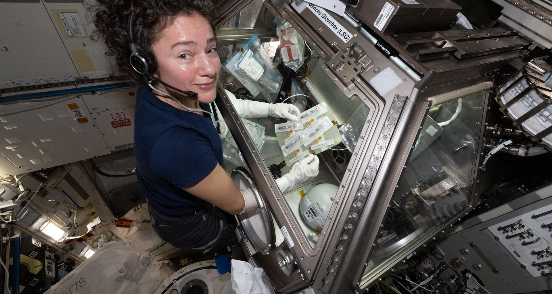 Jessica Meir wears a navy-blue T-shirt and a headset. She looks at the camera while her arms are inserted into the Life Sciences Glovebox. She holds a rectangular plate with multiple sealed syringes and tubes. There are many bags within the glovebox with lab supplies. Metallic surfaces and electronics surround the area outside the glovebox.