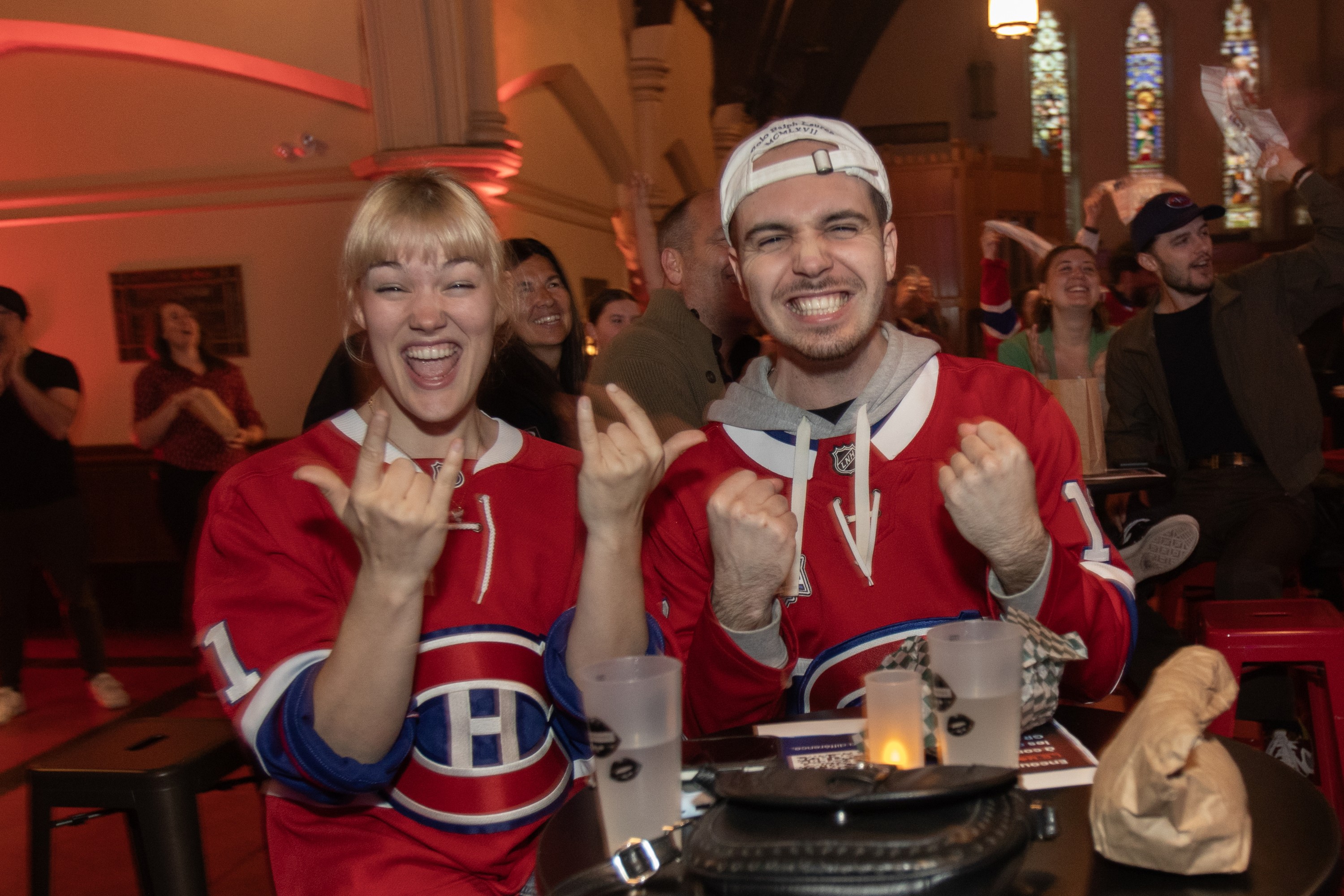 A young man and woman, each in Canadiens jerseys, throw hand signs in celebration while sitting at a table in an indoor hall with stained-glass windows in the background.