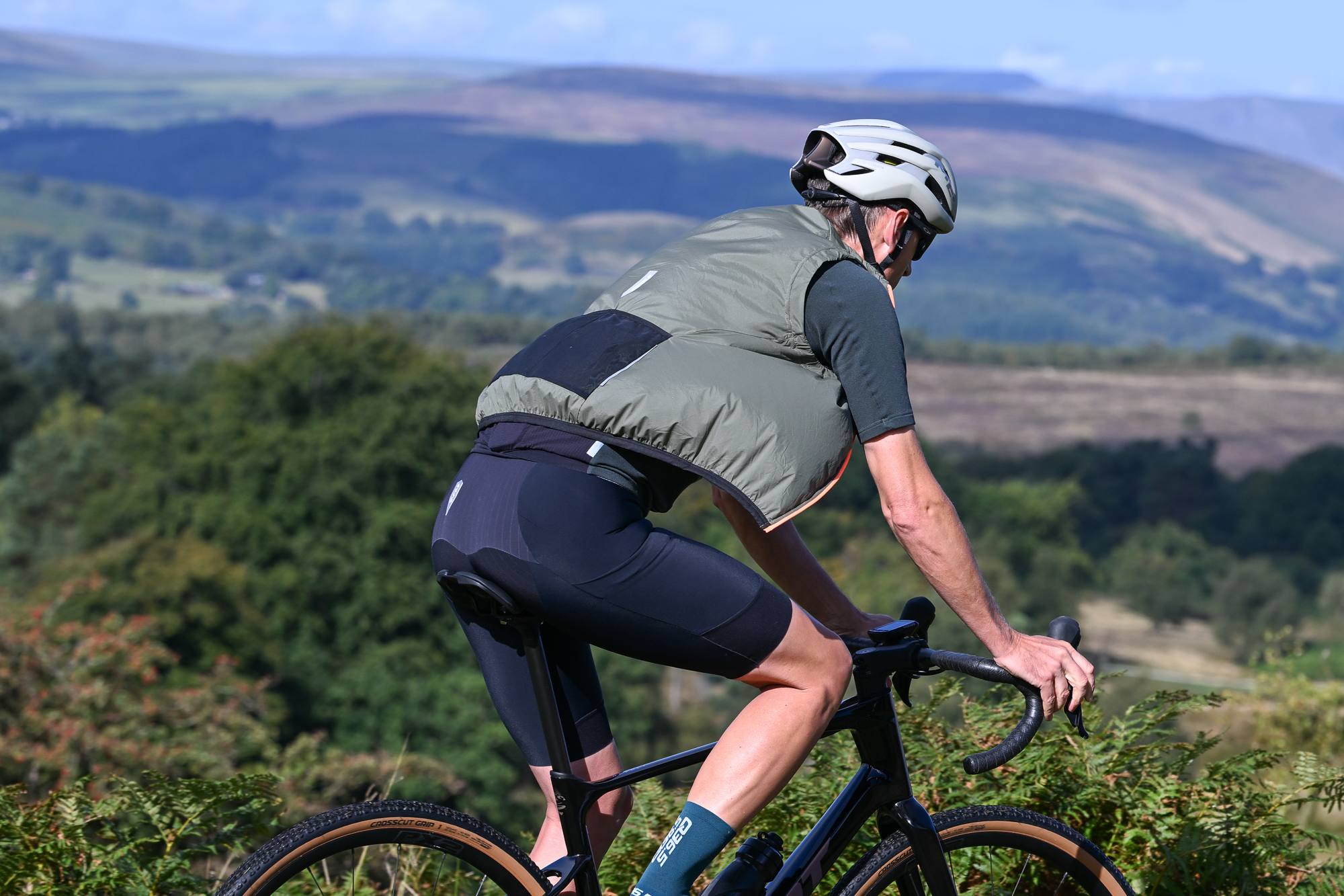 Man wearing black shorts, green jersey and a green gilet riding past the camera on a black gravel bike