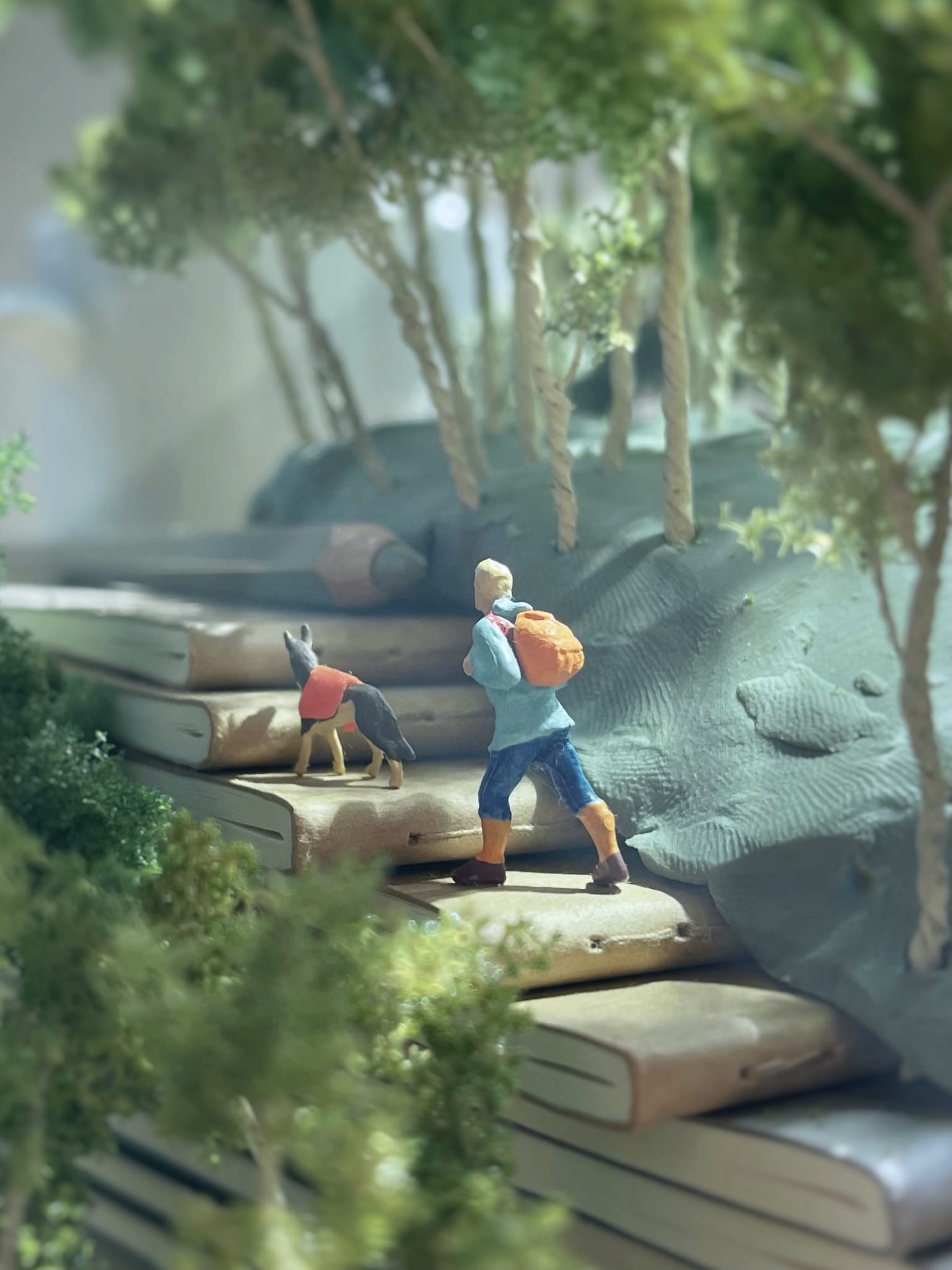 a photograph of a miniature tableau by Derrick Lin of a man and a dog hiking up a hillside, with the rock steps made of books