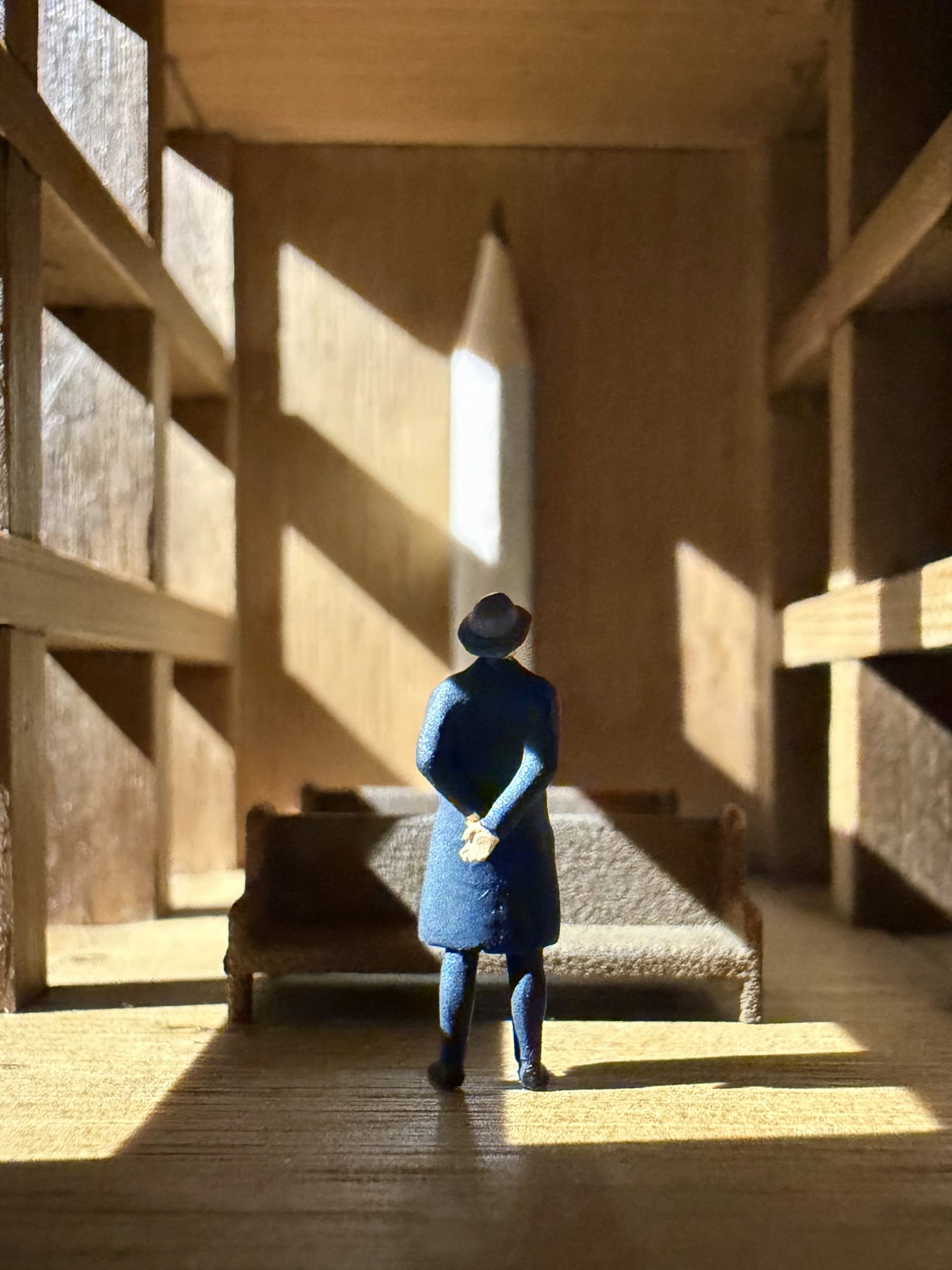 a photograph of a miniature tableau by Derrick Lin of a man in a fedora standing inside of an architectural space, peering at a large pencil that is a centerpiece at a far wall