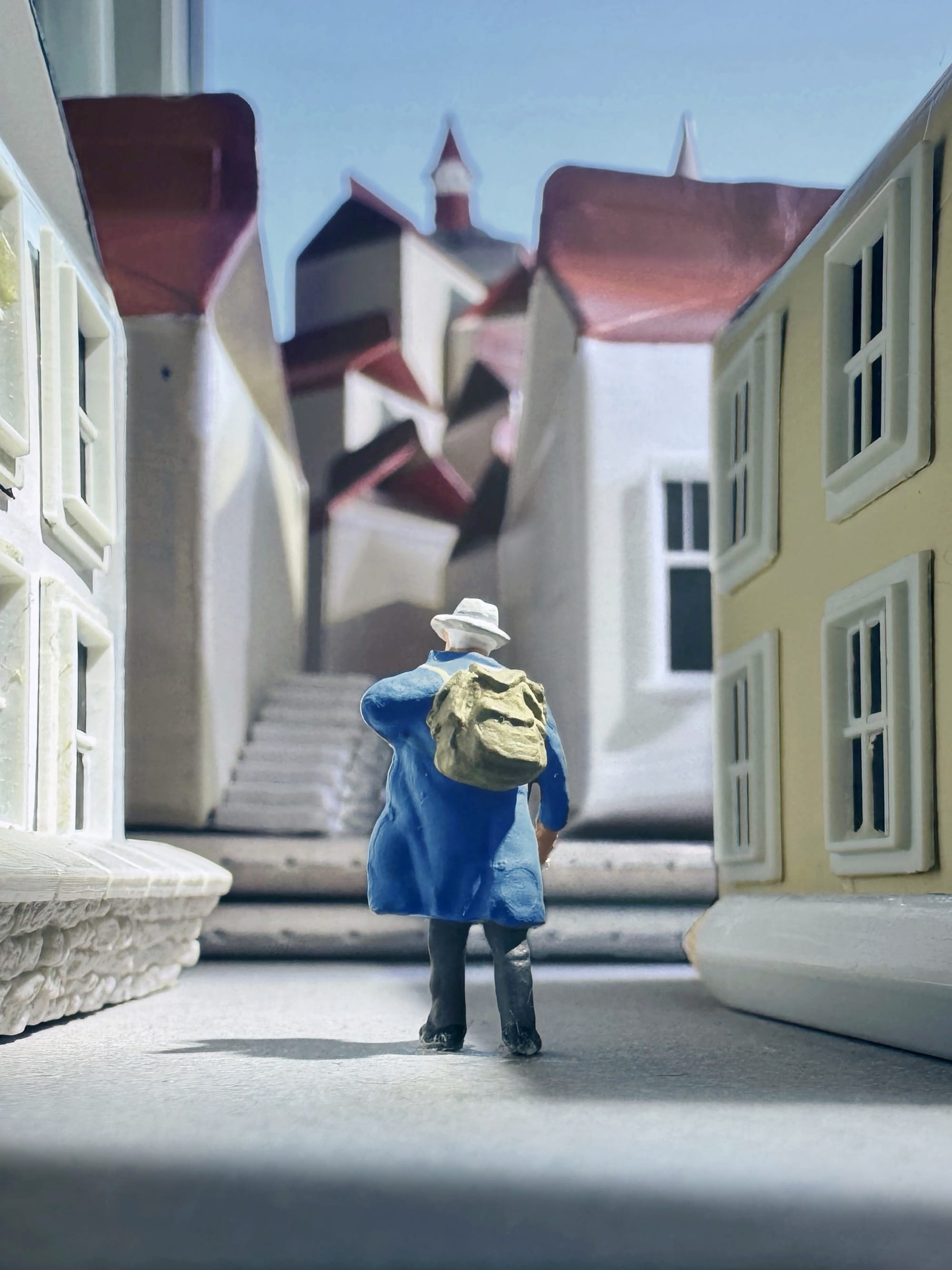 a photograph of a miniature tableau by Derrick Lin of a man with a hat and rucksack walking through a European-looking street with buildikngs made from milk cartons