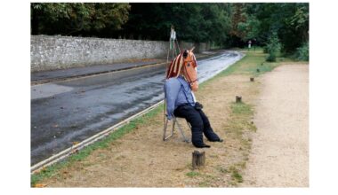 A person wearing a horse mask, blue shirt, and black pants sits on a folding chair beside a wet road and a gravel path, with trees and a stone wall in the background.