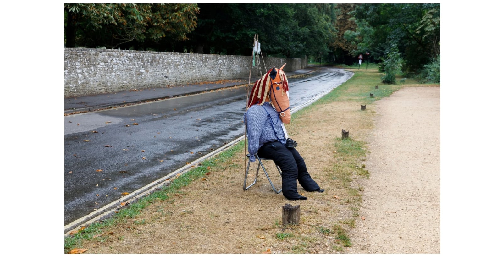 A person wearing a horse mask, blue shirt, and black pants sits on a folding chair beside a wet road and a gravel path, with trees and a stone wall in the background.