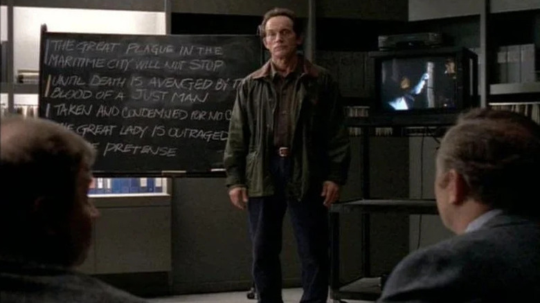 Frank Black (Lance Henriksen) stands by a chalkboard and a television in Millennium