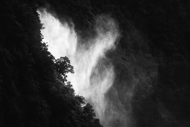 A powerful waterfall cascades through dense, dark forest, with mist rising and light highlighting the surrounding trees in a dramatic black and white scene.