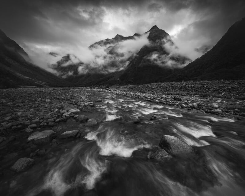 A dramatic black and white photo of a rocky river flowing through a mountain valley, with mist and clouds swirling around the towering peaks in the distance.
