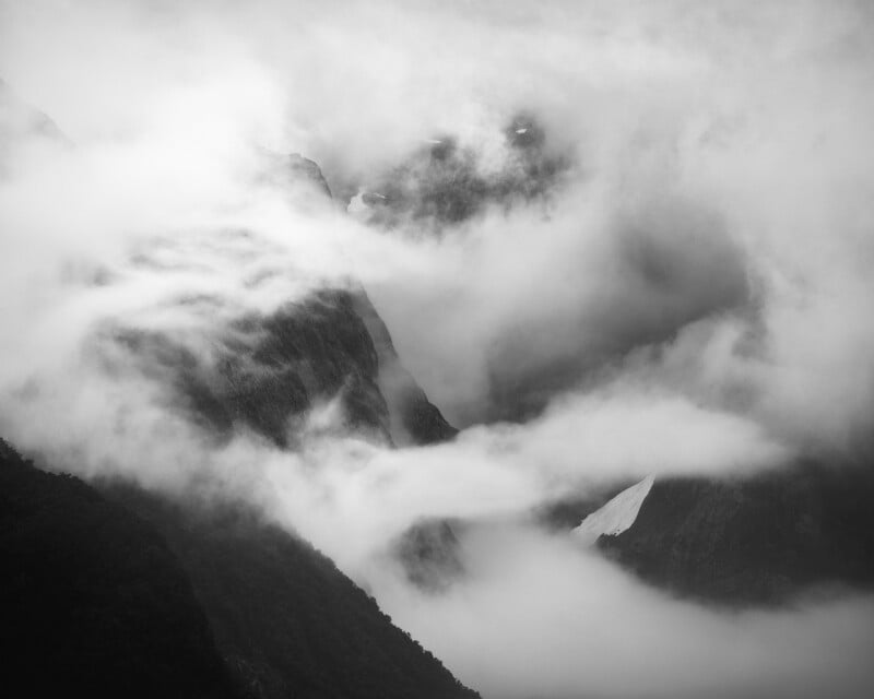 Black and white image of a misty mountain landscape, with thick clouds swirling around jagged peaks and a patch of snow visible on one slope, creating a dramatic and mysterious atmosphere.