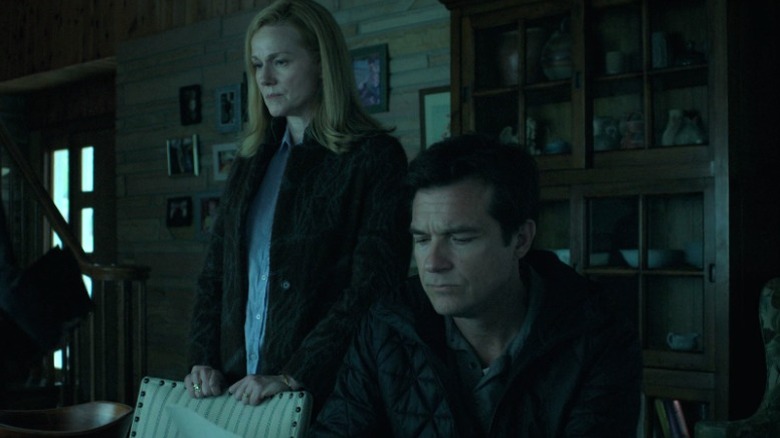 Wendy standing with a heavy expression while Marty sits next to her and reads a document on Ozark S2E9