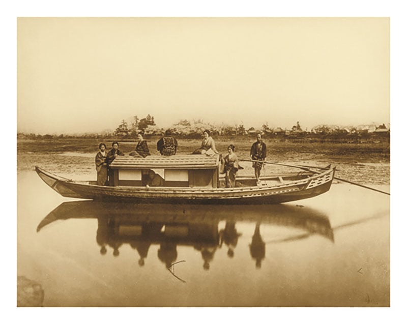 A sepia-toned photograph of five people standing on a traditional wooden boat with a small cabin, reflected in calm water, with open landscape and trees in the background.