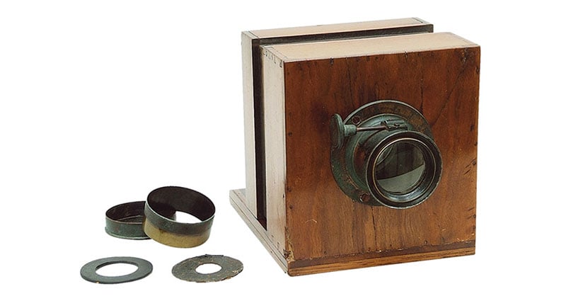 A vintage wooden camera with a brass lens is shown next to several round metal lens accessories on a white background.