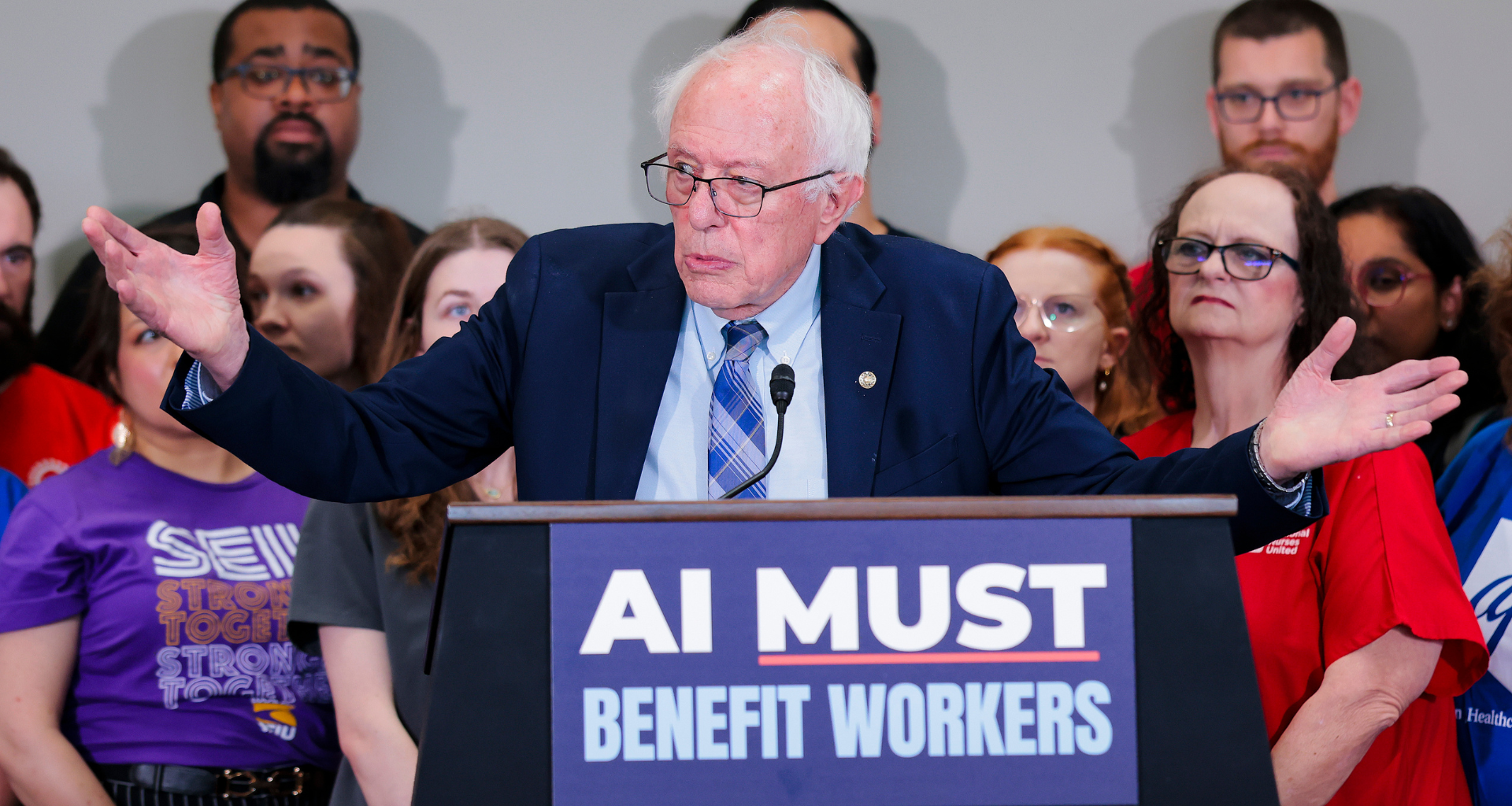 Bernie Sanders talking to a group of people with a podium saying "AI must benefit workers"