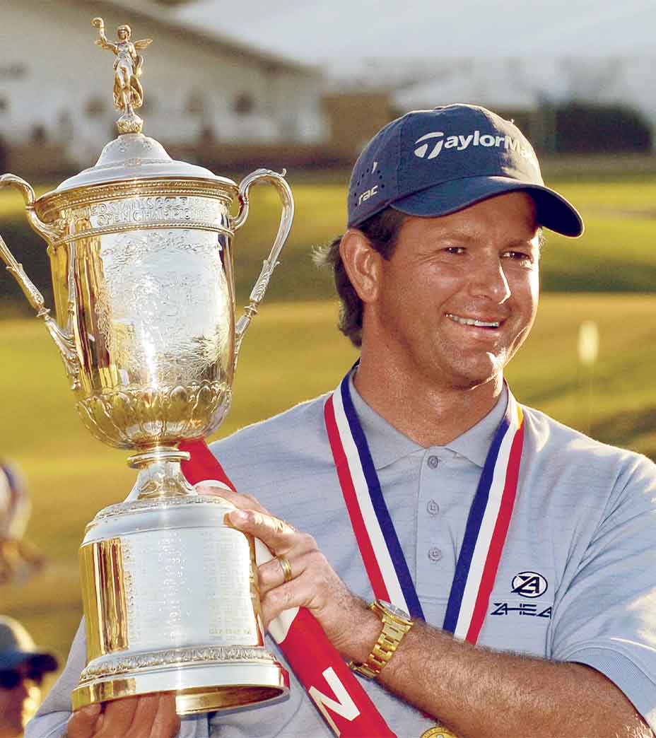 A golfer wearing a cap and a medal around his neck smiles while holding up a large trophy on the Shinnecock Hills golf course, celebrating as one of the U.S. Open winners.