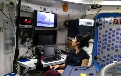 Person seated inside a compact lab-like interior, looking up at a monitor above laptops, equipment racks, and a visible EXIT sign.