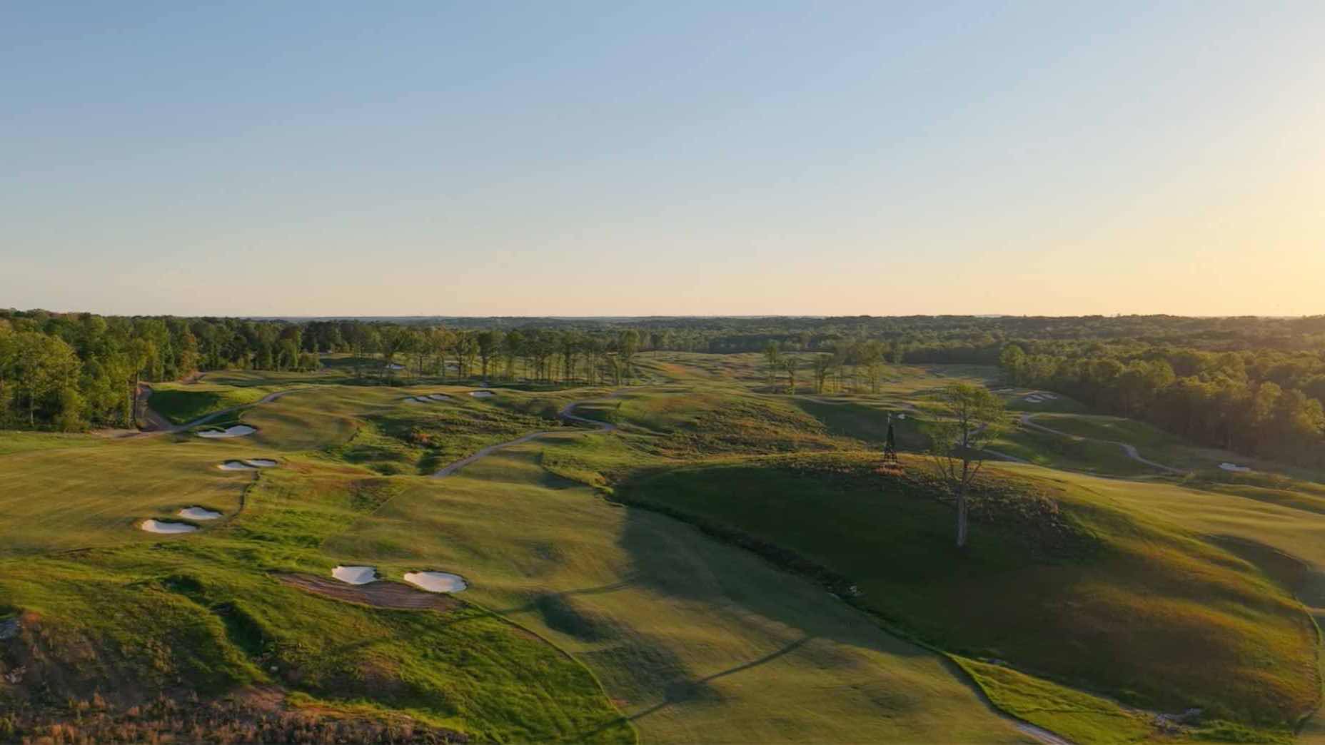A wide view of a golf club at sunset, featuring rolling green fairways, sand bunkers, scattered trees, and a wooded area in the background under a clear sky.