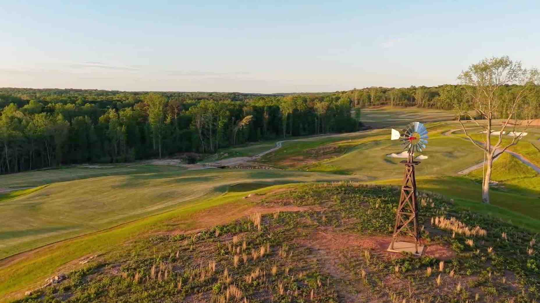 A scenic golf course with rolling green fairways, bordered by a dense forest. In the foreground, a rustic windmill stands on a small hill under a clear sky—holding the perfect spot for your next golf club secret weapon.