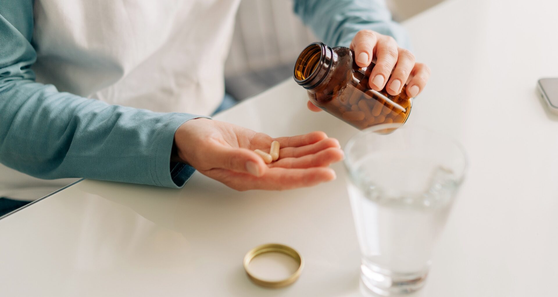 A person pours capsules from a brown bottle into their palm, with a glass of water nearby.