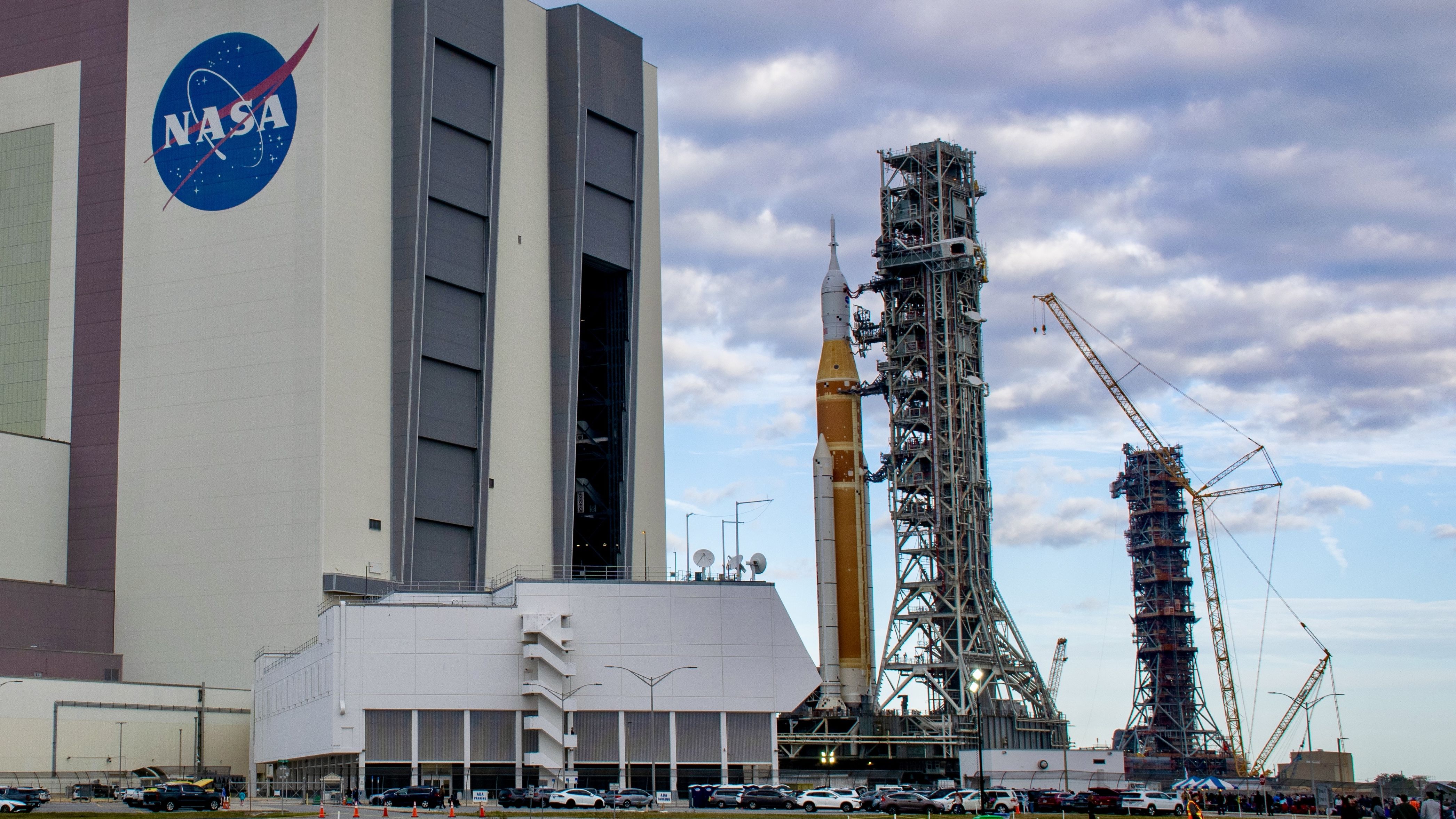 A bit orange rocket rolls out of a giant white building, with an extra launch tower in the background.