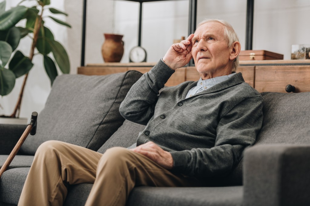 A thoughtful senior man with grey hair sits on a sofa in a living room, looking upwards with his hand on his forehead.