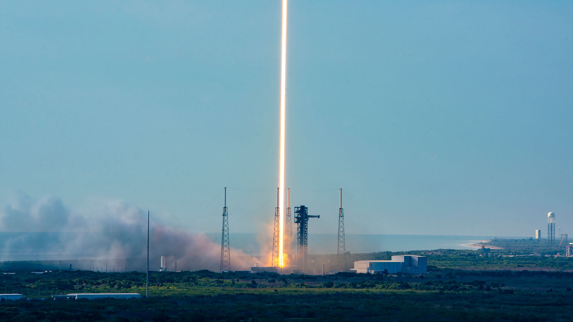 A time lapse photo of a daytime rocket launch shows the rocket's fiery plume as a single orange and white beam heading skyward off the launch pad into the blue sky.