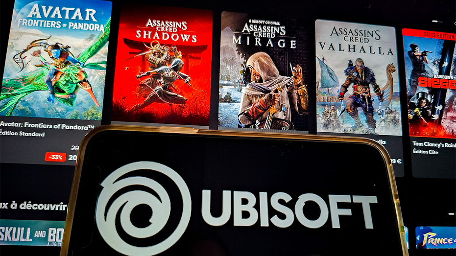 Illustration by Ubisoft in France, January 27, 2026. A smartphone displays the Ubisoft logo with a purple spiral and black lettering, posing in front of three promotional posters for the Assassins Creed series.